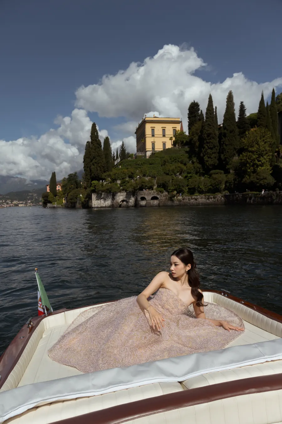 Karin reclining on a wooden boat on the lake, a villa and cypress trees behind